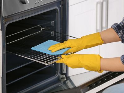 person wearing yellow rubber gloves with a blue spong cloth wiping an oven shelf
