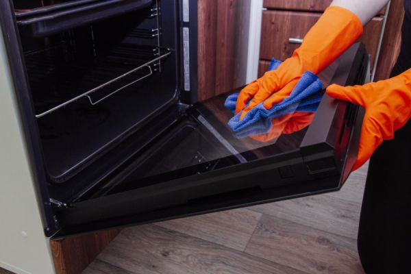 hand in orange gloves cleaning the door of an oven with a sponge
